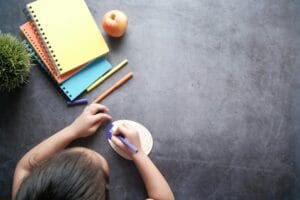 Top view of a child drawing on a textured surface with colorful notebooks and an apple nearby.